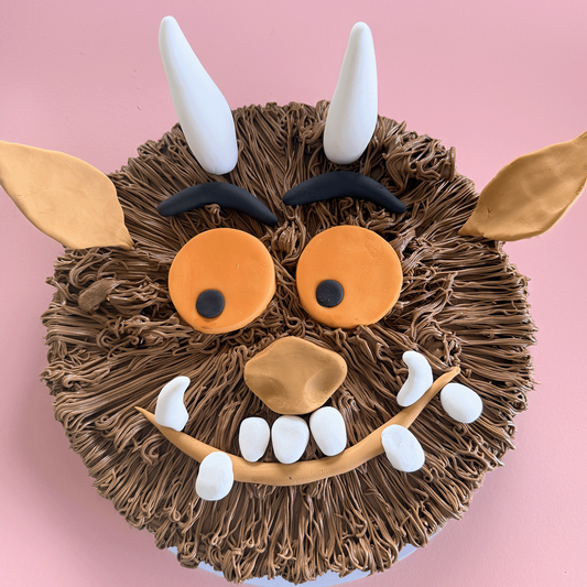 A round cake decorated to look like the Gruffalo, featuring brown textured frosting hair, orange eyes, white horns, and a smiling mouth, set against a solid pink background.