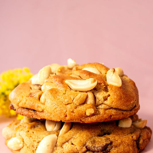 Two golden-brown cookies stacked on top of each other, topped with peanuts, set on a soft pink background with small yellow flowers at the side.