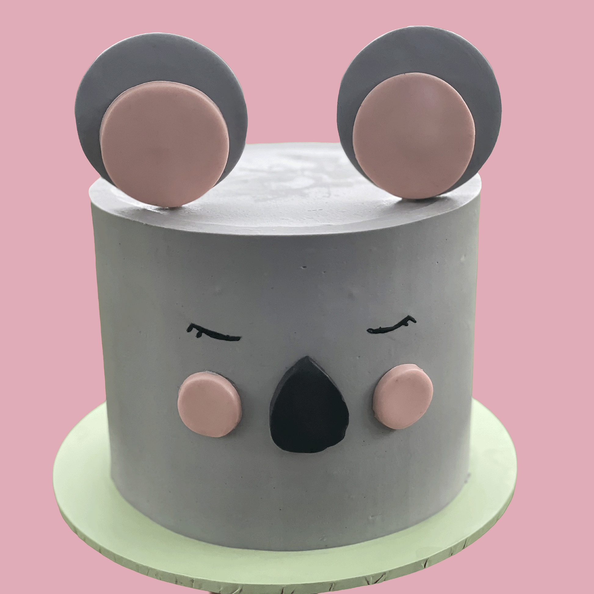 A round gray frosted cake decorated to look like a koala face, with pink inner ears, black closed eyes and nose, and a pale green cake stand against a solid pink background.