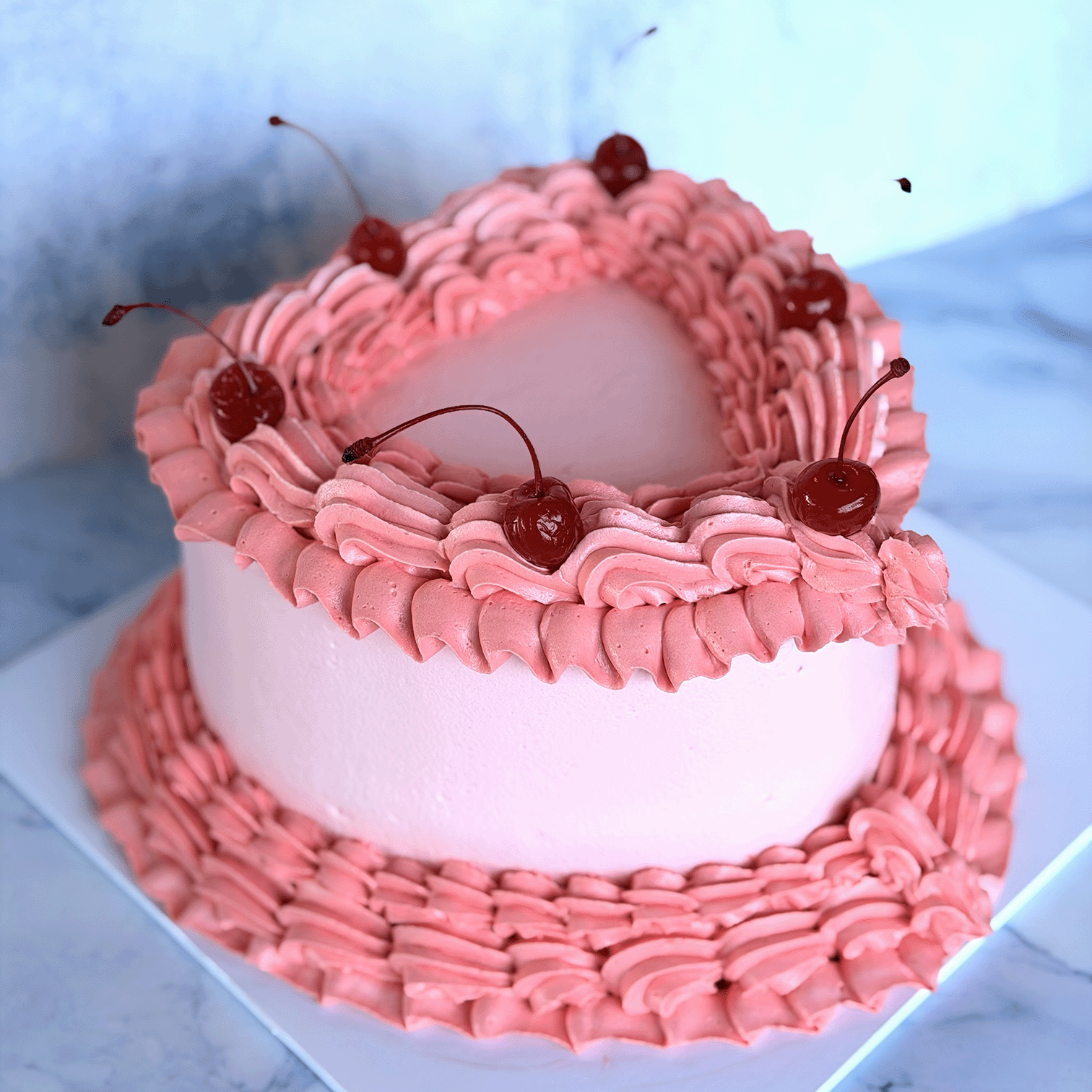 A pink frosted heart-shaped cake decorated with piped borders, cherries on top, and a matching pink frosting base, shown on a white board against a pale blue background.